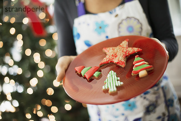 Eine Frau  die eine Schürze trägt und einen Teller mit organisch dekorierten Weihnachtsplätzchen hält.