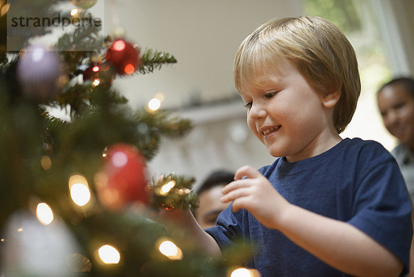 Ein kleiner Junge hält Weihnachtsschmuck in der Hand und legt ihn auf den Weihnachtsbaum.