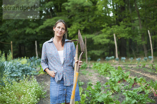 Eine Frau  die in einem biologischen Garten arbeitet und zwischen den Gemüsekulturen steht. Sie hält einen Spaten und eine Hacke in der Hand.