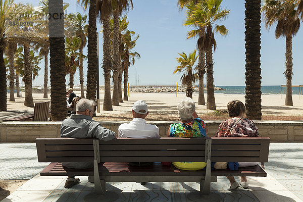 Rückansicht von Touristen die sich auf einer Bank ausruhen  Ausblick auf Palmen und Strand  Salou  Katalonien  Spanien