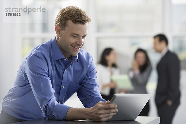 Junge Berufstätige bei der Arbeit. Ein Mann in einem Hemd mit offenem Hals  der ein digitales Tablet benutzt. Eine Gruppe von Männern und Frauen im Hintergrund.