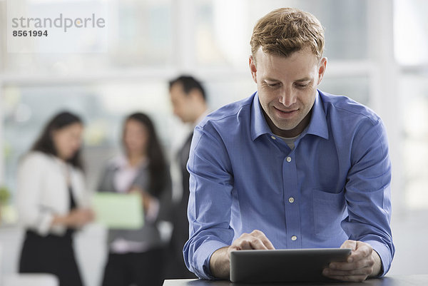 Junge Berufstätige bei der Arbeit. Ein Mann in einem Hemd mit offenem Hals  der ein digitales Tablet benutzt. Eine Gruppe von Männern und Frauen im Hintergrund.