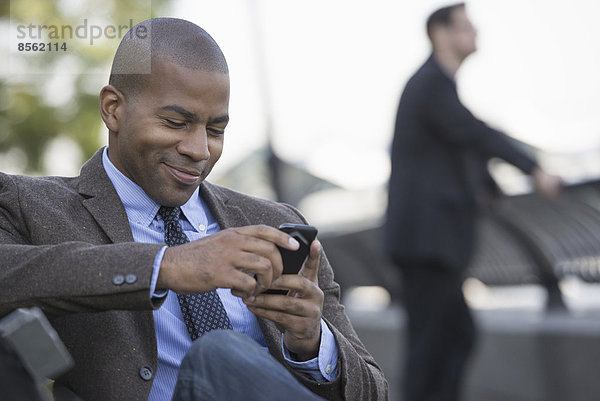 Geschäftsleute in der Stadt. Unterwegs in Kontakt bleiben. Ein Mann sitzt mit seinem Smartphone. Ein Mann im Hintergrund.