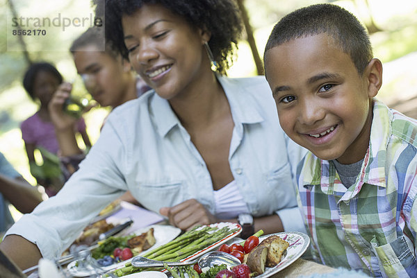 Ein Familienpicknick im Schatten hoher Bäume. Eltern und Kinder bedienen sich an frischem Obst und Gemüse.