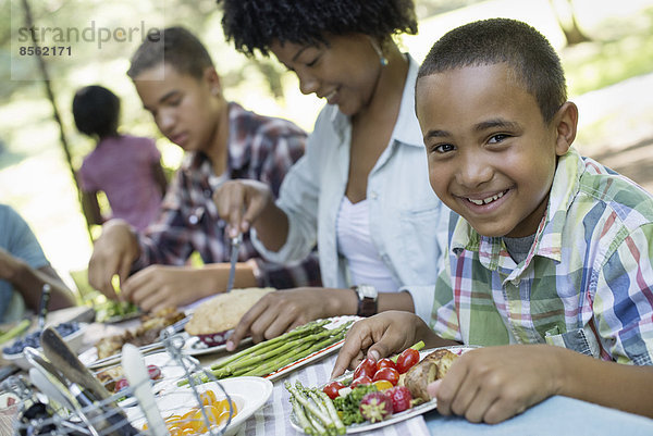 Ein Familienpicknick im Schatten hoher Bäume. Eltern und Kinder bedienen sich an frischem Obst und Gemüse.