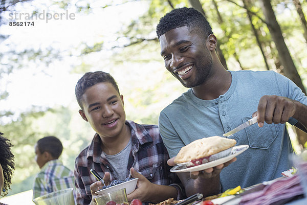 Ein Familienpicknick im Schatten hoher Bäume. Eltern und Kinder bedienen sich an frischem Obst und Gemüse.