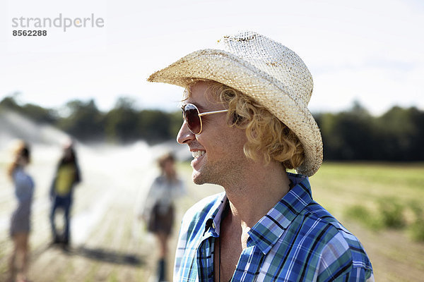 Ein junger Mann mit Strohhut und Sonnenbrille. Bewässerungssprinkler auf dem Feld.
