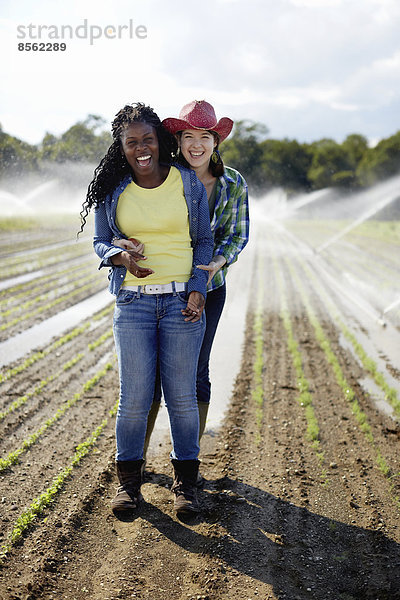 Zwei junge Frauen stehen auf einem Feld mit kleinen Setzlingen  auf dem die Bewässerungssprinkler den Boden besprühen.