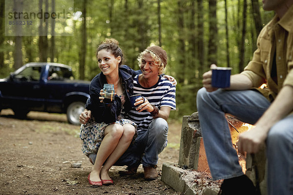 Ein Campingplatz in den Wäldern. Drei Personen sitzen in der Abenddämmerung um ein Lagerfeuer herum.