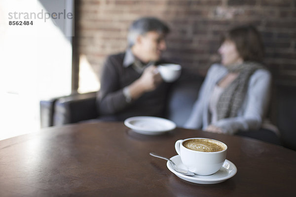 Zwei Personen sitzen in einem Café. Ein Mann hält ein weißes Porzellan in der Hand und trinkt. Sitzt neben einer Frau. Ein Tisch mit einer großen vollen Tasse Cappuccino-Kaffee.