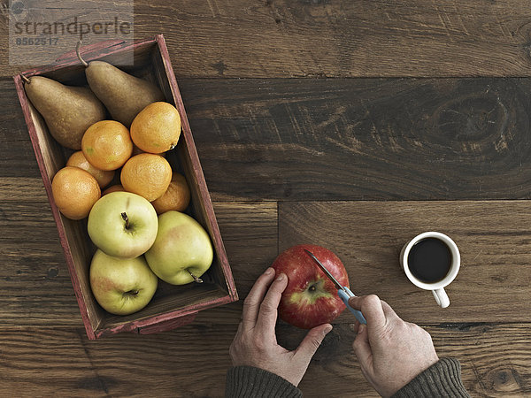 Eine Tischplatte aus Holz mit verschiedenen Holzfarben und Maserungen. Eine Kiste mit frischen Früchten  Birnen und Orangen. Eine Person  die mit einem Messer einen Apfel hackt.