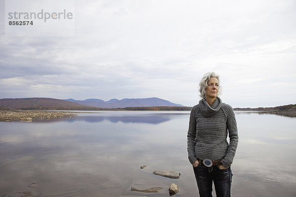 Eine Frau  die am Ufer eines ruhigen Sees über das Wasser schaut.