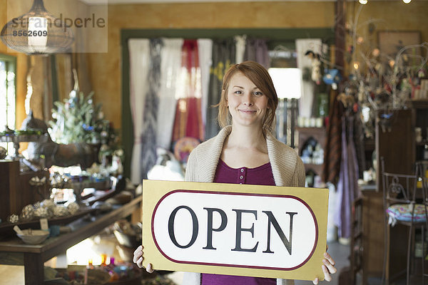 Eine Frau steht in einem Antiquitätengeschäft und hält ein OPEN-Schild. Überall um sie herum sind Waren ausgestellt.