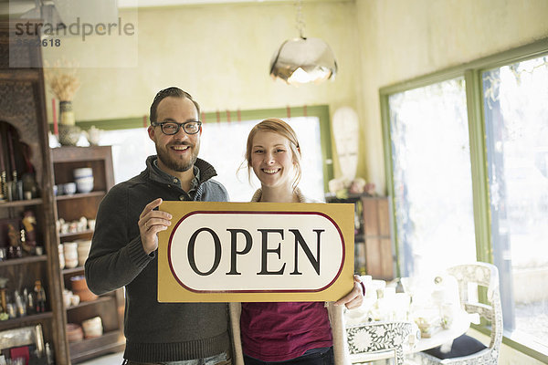 Zwei Personen stehen in einem Laden voller antiker Gegenstände  ein Ehepaar führt ein Geschäft. Sie halten ein großes Schild mit der Aufschrift OPEN.