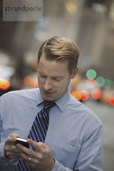 Ein Mann in Hemd und Krawatte  der sein Handy überprüft  steht auf einer belebten Straße.