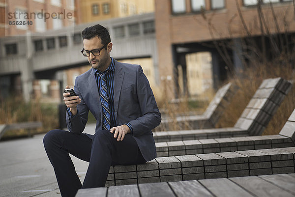 Ein Mann in formeller Jacke und Krawatte  der auf einer Bank vor einem städtischen Gebäude sitzt und sein Telefon auf Nachrichten überprüft.