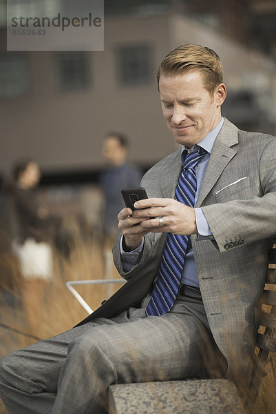 Ein Mann  der auf einer Bank vor einem großen Gebäude sitzt und auf den Bildschirm eines Mobiltelefons oder Handys schaut.