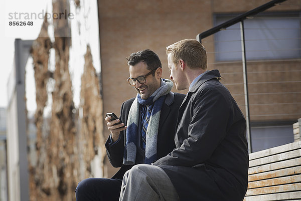 Zwei Männer sitzen auf einer Bank vor einem großen Gebäude in der Stadt. Einer überprüft sein Mobiltelefon.