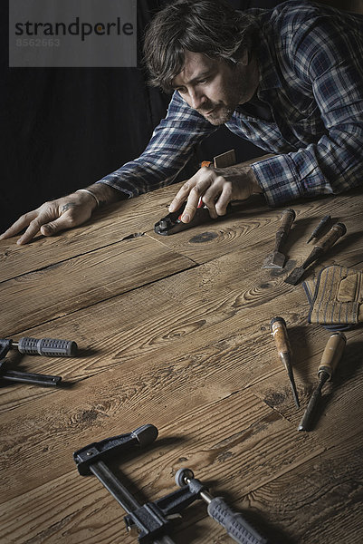 Ein Mann  der in einer wiederaufgearbeiteten Holzlagerhalle arbeitet. Halten von Werkzeugen und Schleifen von verästelten und unebenen Holzstücken.
