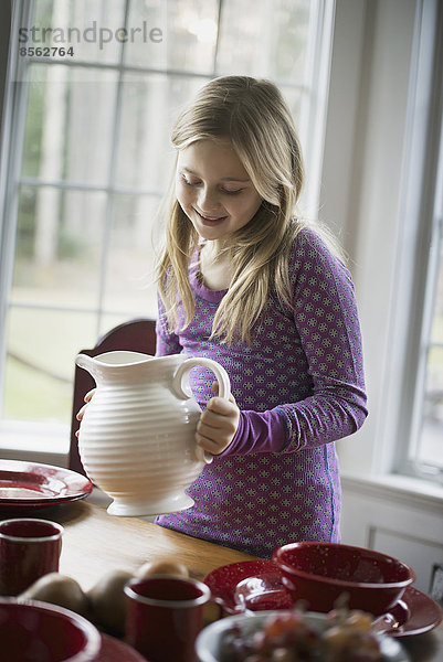 Kinder in einem Familienhaus. Ein Mädchen hält einen weißen Tonkrug. Mit Geschirr gedeckter Tisch.