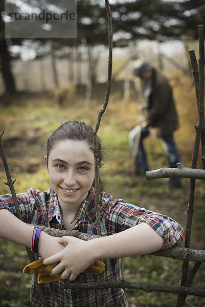 Ein junges Mädchen lehnt an einem Zaun  auf einem Biohof. Im Hintergrund ein Mann  der den Boden umgräbt.