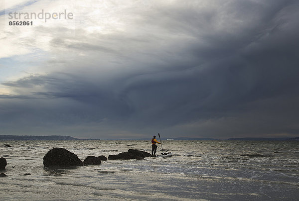 Ein Mann steht auf einem Felsen mitten im Puget Sound  neben ihm schwimmt sein Seekajak.