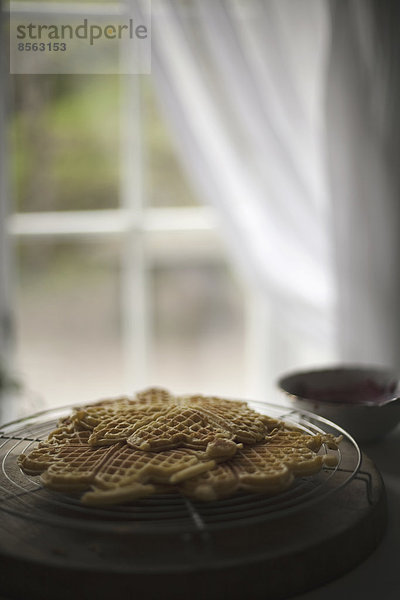 Eine Waffelschale auf einer Tischplatte. Von einem Fenster zurück drapierte Gardinen.