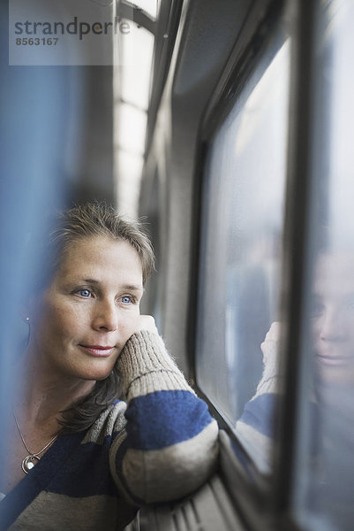 Eine Frau sitzt an einem Fensterplatz in einem Zugwaggon  den Kopf auf die Hand gestützt. Sie schaut in die Ferne.