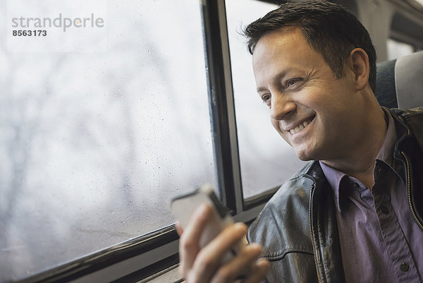 Ein reifer Mann  der an einem Fensterplatz in einem Zug sitzt und sein Mobiltelefon in der Hand hält. Er lächelt und schaut in die Ferne.
