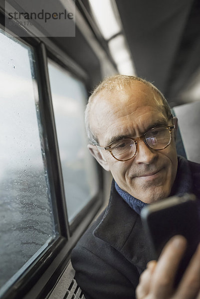 Ein reifer Mann  der in einem Zugwaggon am Fenster sitzt  sein Mobiltelefon benutzt und während der Fahrt in Kontakt bleibt.