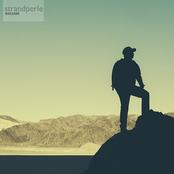 Silhouette eines Wanderers mit Rucksack  der im Death Valley National Park mit Blick auf Berge und Wüste eine Pause einlegt.