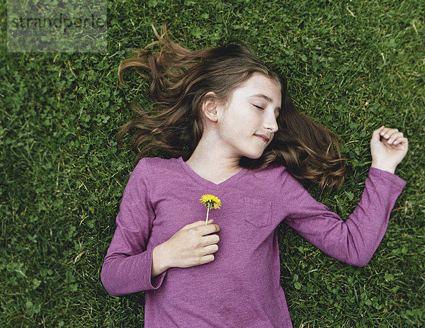 Ein zehnjähriges Mädchen  das mit geschlossenen Augen im Gras liegt und eine Löwenzahnblume hält.