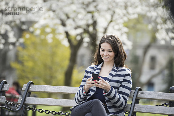 Draußen in der Stadt im Frühling. Park von New York City. Weiße Blüte an den Bäumen. Eine Frau sitzt auf einer Bank und hält ihr Mobiltelefon.