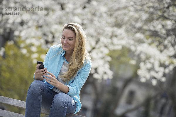 Draußen in der Stadt im Frühling. Park von New York City. Weiße Blüte an den Bäumen. Eine Frau sitzt auf einer Bank und hält ihr Mobiltelefon.