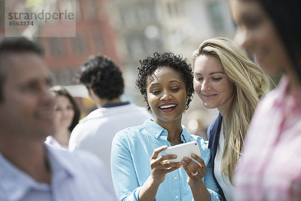 Menschen im Frühling in der Stadt im Freien. New York City. Eine Gruppe von Männern und Frauen  zwei Frauen mit Blick auf ein Mobiltelefon.