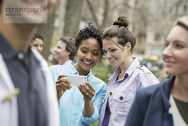 Menschen im Frühling in der Stadt im Freien. Der New Yorker Stadtpark. Zwei Frauen in einer Gruppe von Freunden  die auf ein Handy schauen und lächeln.