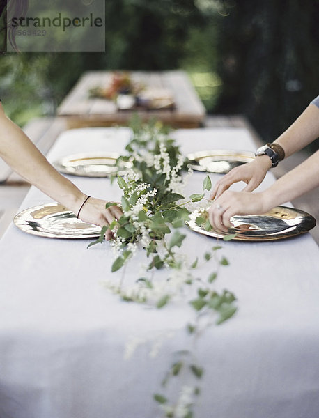 Zwei Personen lehnen sich über einen Tisch  der außen mit einem weißen Tuch und einer Tischdekoration aus Laub in der Mitte gedeckt ist. Platzieren von Besteck und Tellern auf einer Tischplatte.