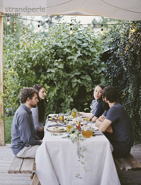 Vier Personen saßen an einem Tisch im Garten. Platzieren Sie Gedecke und Dekorationen auf einer weißen Tischdecke. Zwei Männer und zwei Frauen.