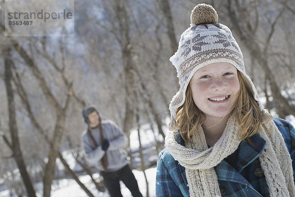 Winterlandschaft mit Schnee auf dem Boden. Ein junges Mädchen mit Pudelmütze und Schal im Freien. Im Hintergrund ein Mann.