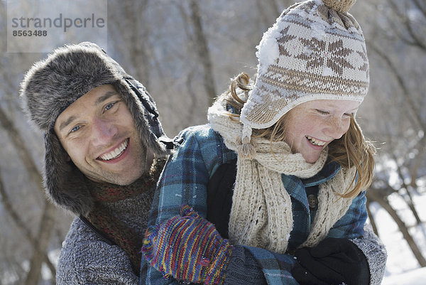 Winterlandschaft mit Schnee auf dem Boden. Ein junges Mädchen mit Pudelmütze und Schal und ein Mann  der sie umarmt.