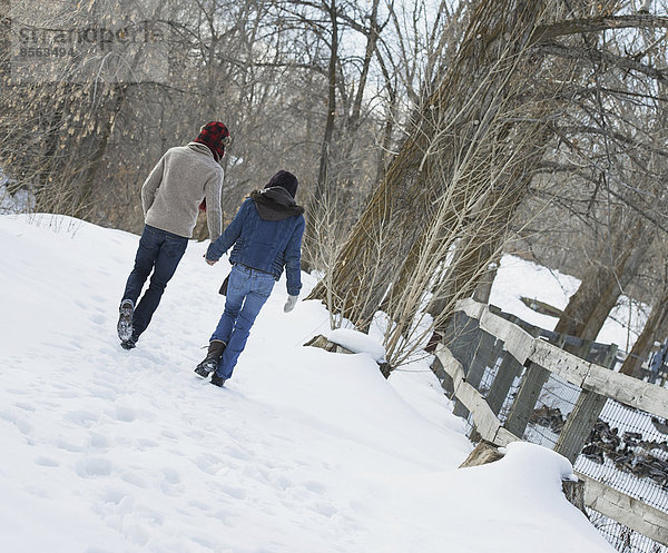 Winterlandschaft mit Schnee auf dem Boden. Ein Paar geht Hand in Hand einen Weg entlang.