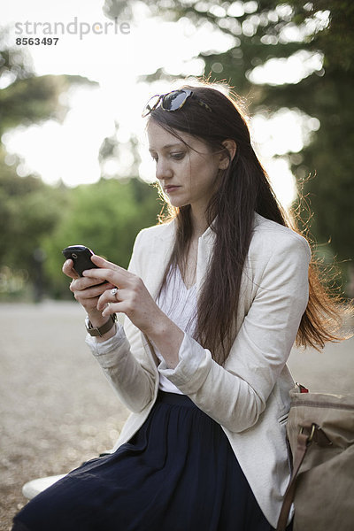 Eine Frau in einer cremefarbenen Jacke  die in einem Stadtpark sitzt und auf ihr Handy schaut.