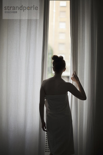 Eine junge Frau mit hochgestecktem Haar  ein Badetuch tragend  schaut durch lange Vorhänge auf ein Fenster.