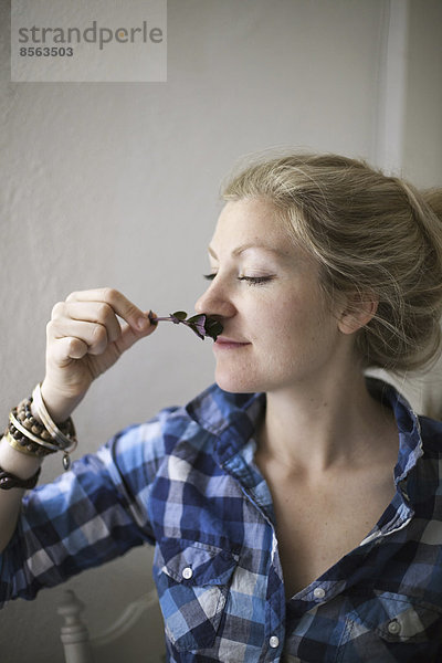 Eine junge Frau mit blonden Haaren  die eine aromatische Pflanze oder Kräuterblüte an ihre Nase hält und das Aroma einatmet.