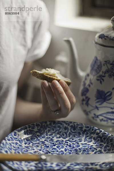 Ein Frühstückstisch mit einer hohen Kaffeekanne und einem blau-weißen Porzellanteller. Eine Frau sitzt mit einem Stück Butterbrot in der Hand.