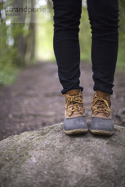 Eine Frau in Wanderstiefeln und Jeans  die auf einem Stein neben einem Waldweg steht.
