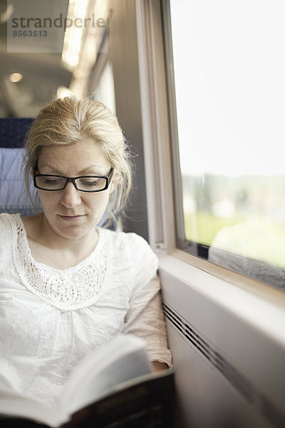 Eine Frau sitzt an einem Zugfenster und liest ein Buch.