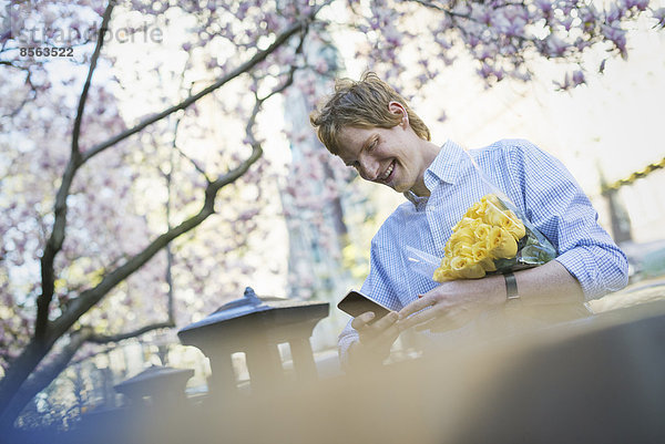 Stadtleben. Ein junger Mann im Frühling im Park  der ein Mobiltelefon benutzt. Er hält einen Strauß gelber Rosen in der Hand.