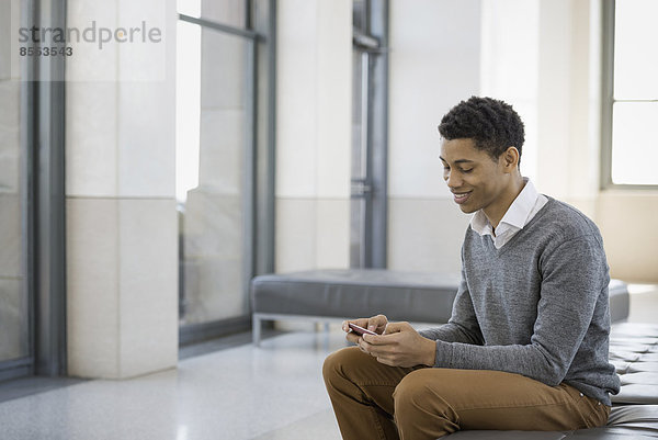 Urbaner Lebensstil. Ein junger Mann sitzt in einer Lobby  auf einer Sitzbank. Er benutzt sein Mobiltelefon.