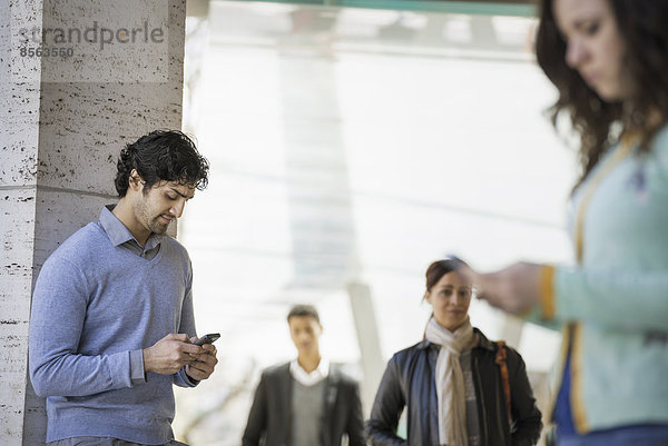 Urbaner Lebensstil. Drei Personen auf dem Bürgersteig  ein Mann und eine junge Frau  die ihre Mobiltelefone benutzen  und zwei Personen  die auf dem Bürgersteig gehen.